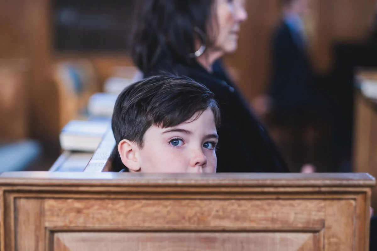 A young boy with blue eyes looks over the wooden back of a church pew during a wedding ceremony.