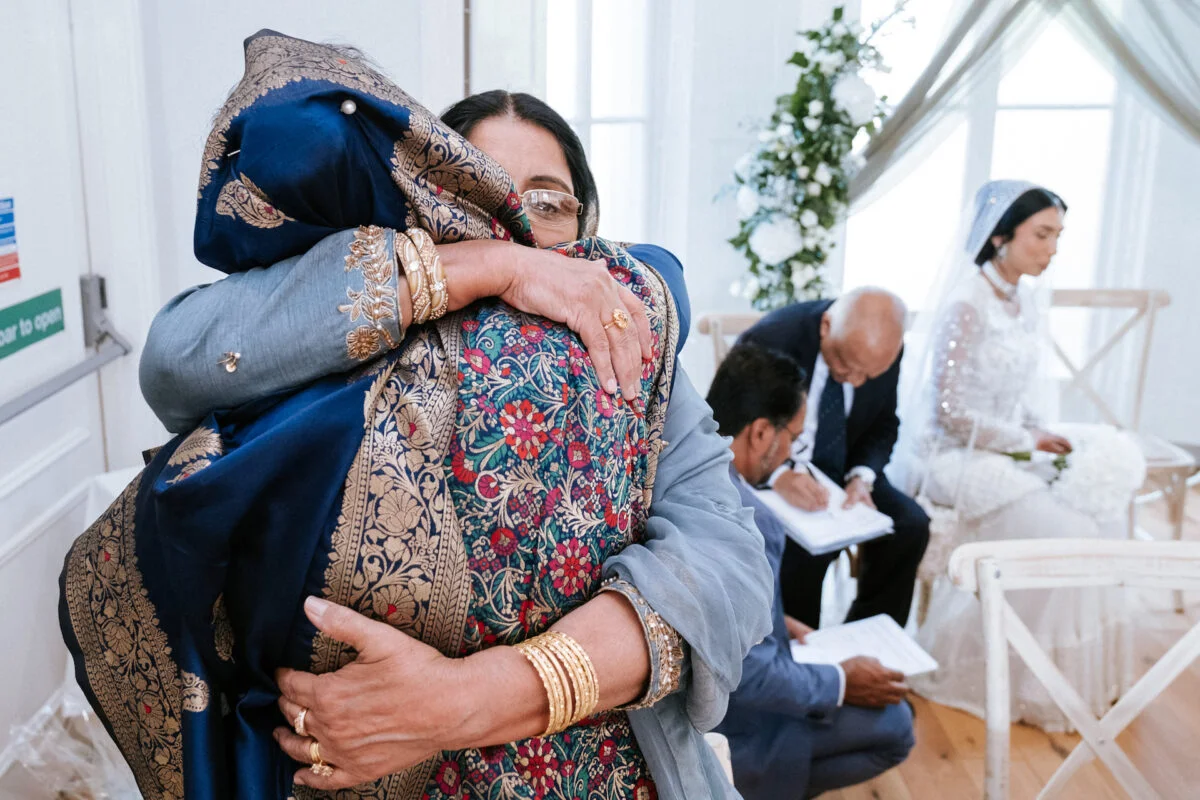 An emotional family member shares a tight embrace during a wedding ceremony while the couple signs the register in the background.
