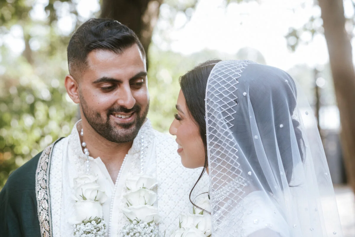 A newly married couple share a loving glance outdoors during their wedding ceremony, with the groom wearing a traditional embroidered outfit and white rose garlands, and the bride wearing an embellished veil.