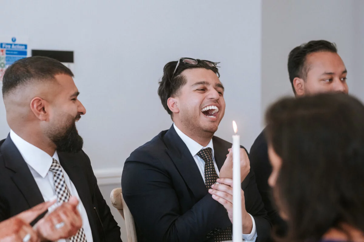 A groom and his groomsmen share a joyful belly laugh together during the wedding speeches, seated by a glowing candle.