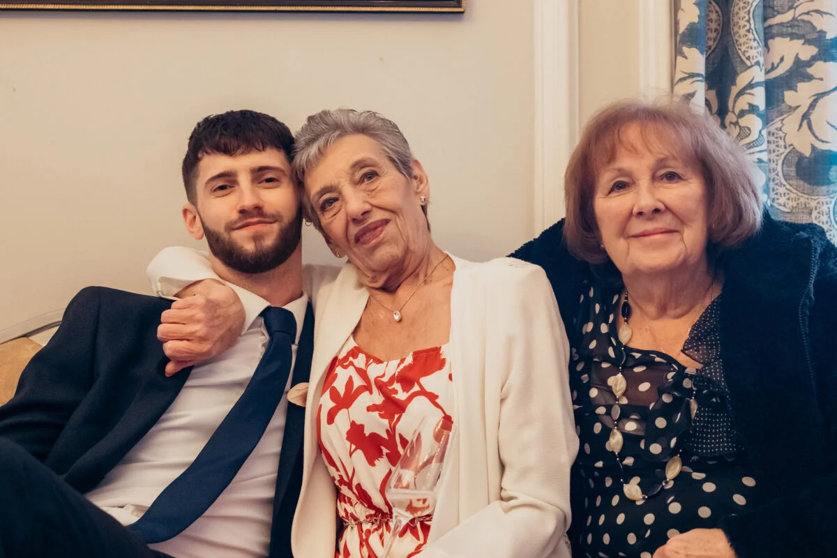 A young groom with a beard and dark suit sits closely beside two older family members indoors, smiling warmly for the camera.