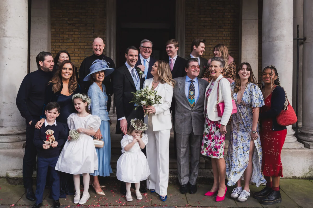 A large, joyful group of wedding guests and family members gather outside a historic stone-columned venue, smiling and laughing together.