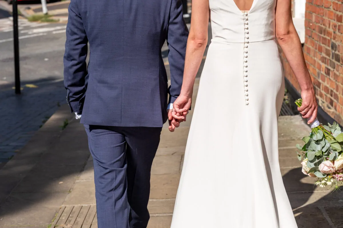 A newlywed couple walk hand in hand down a sunlit city street, with the groom wearing a navy suit and the bride in a button-back crepe gown holding a bouquet.
