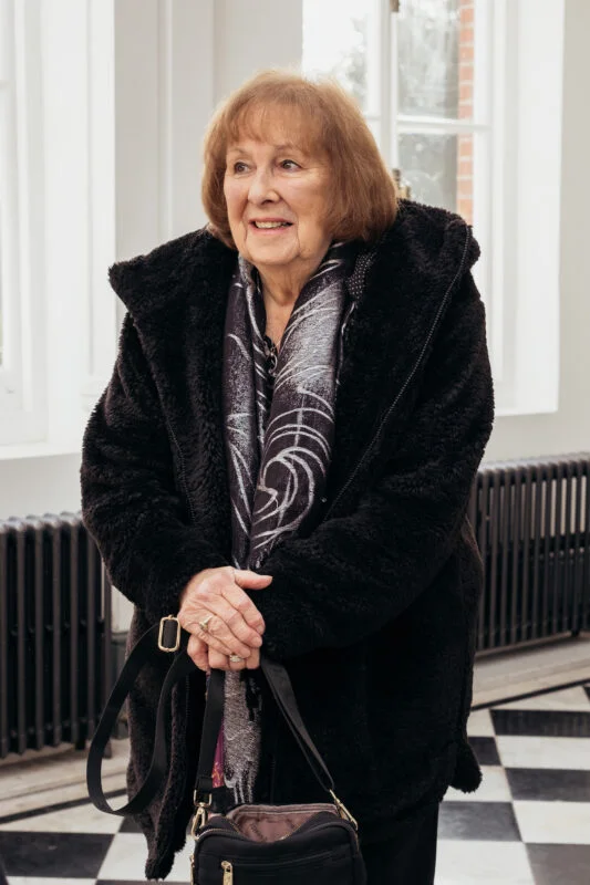 An elderly female wedding guest with short blonde hair smiles warmly indoors while wearing a black fuzzy coat and patterned scarf.