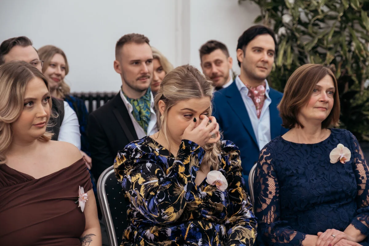 An emotional wedding guest wiping away a tear while seated amongst a gathered crowd of onlookers during the ceremony.