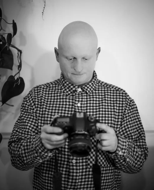 A bald man wearing a checked shirt looks down intently at a digital camera held in his hands in this black and white documentary portrait.