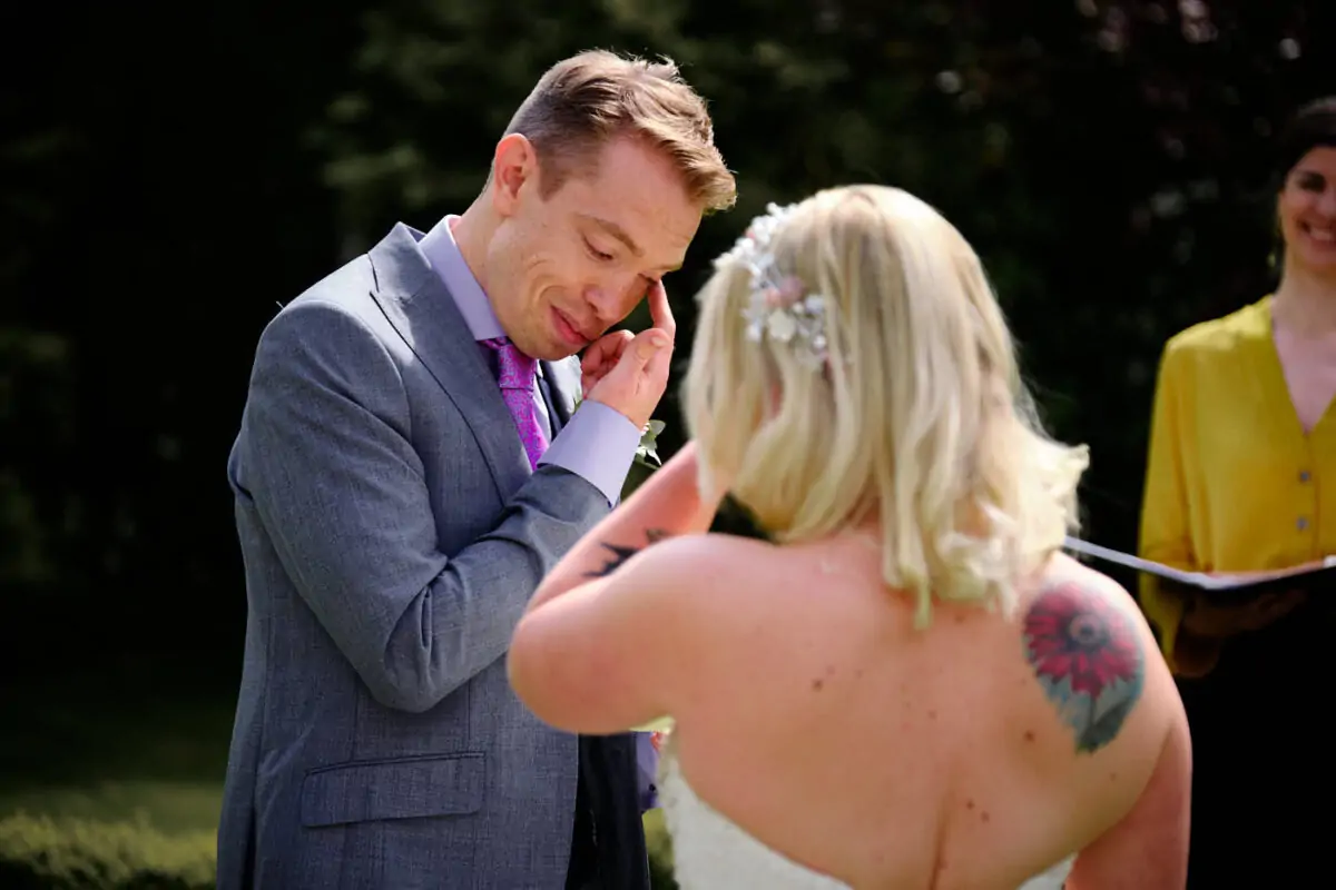 An emotional groom wipes away tears during an outdoor wedding ceremony while standing opposite his bride.