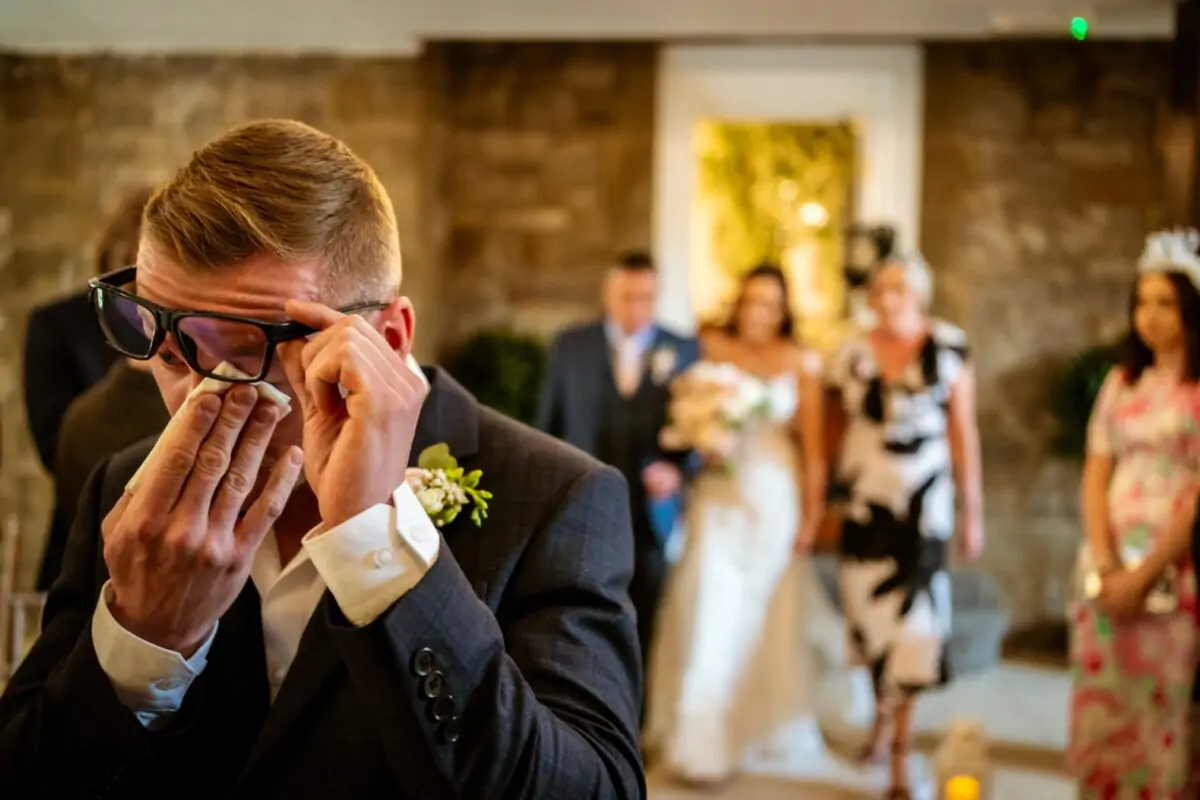 An emotional groom wipes away tears with a tissue as he watches his bride walk down the aisle during a rustic barn wedding ceremony.