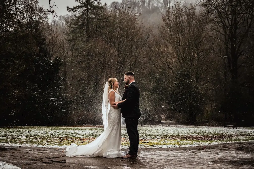 A newlywed couple embraces lovingly outdoors amidst a wintry snowfall against a moody woodland backdrop.