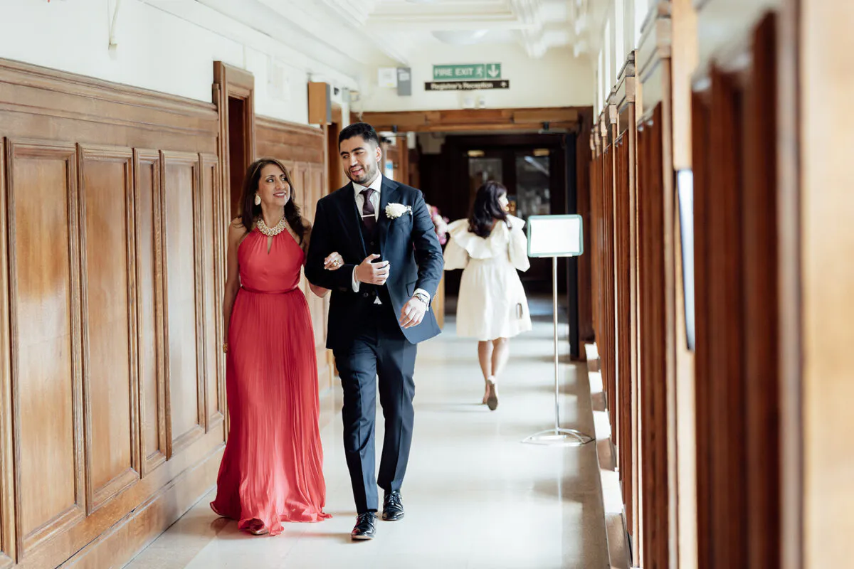 A groom and a woman in a red dress walk arm in arm down a wood-panelled hallway during a wedding celebration.