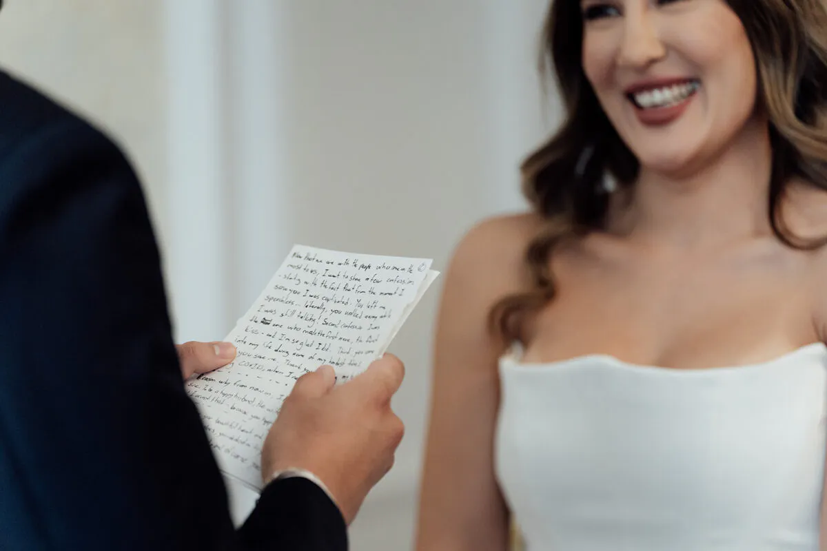 A groom reads handwritten vows from a letter while his smiling bride looks on during the wedding ceremony.