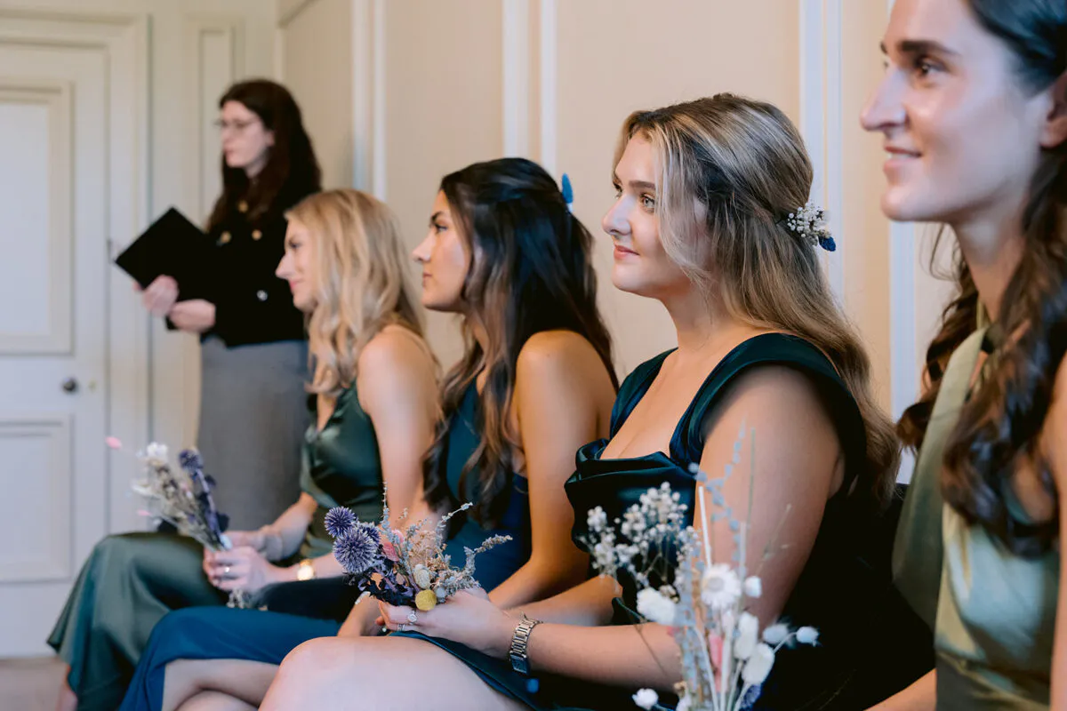 A row of bridesmaids in elegant emerald green satin dresses sit together holding small dried flower bouquets during a wedding ceremony.