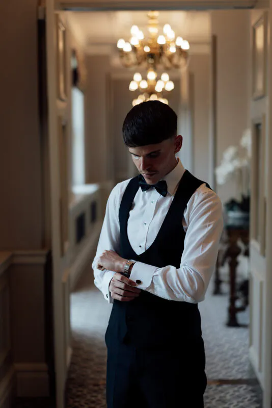 A groom in a classic black tuxedo and bow tie fastens his cufflinks inside an elegant hallway lit by a sparkling chandelier.