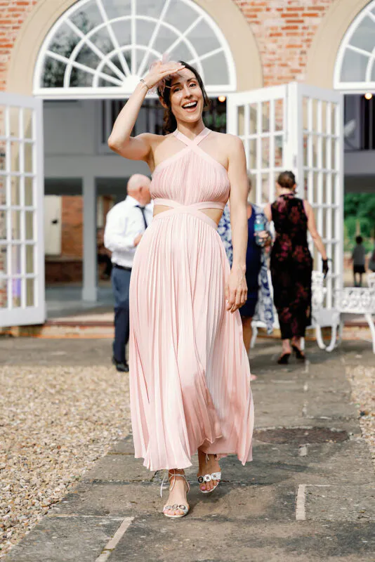 A joyful wedding guest in a pink halter neck pleated dress smiles while walking outdoors on a sunny day.