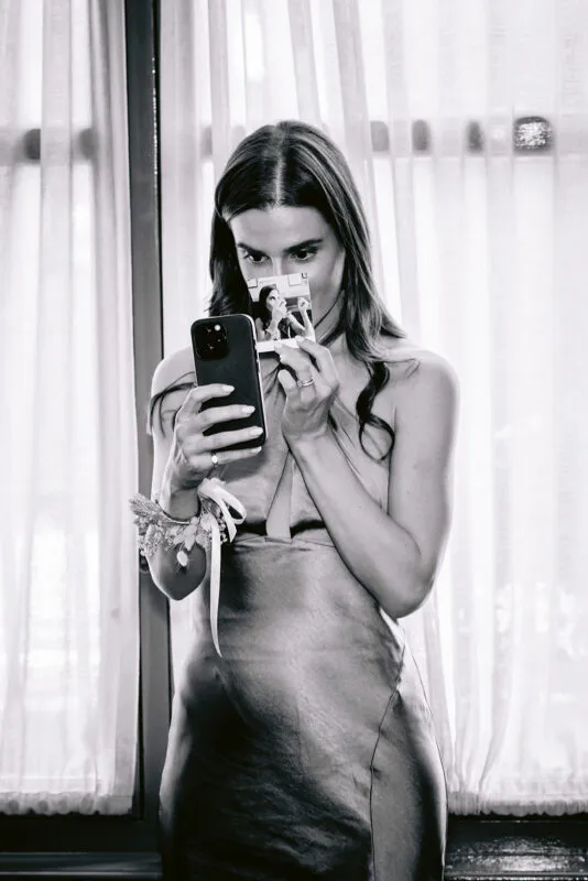 A black and white candid shot of a bridesmaid in a silk slip dress holding a smartphone and looking at a polaroid photo while standing by a window during morning preparations.