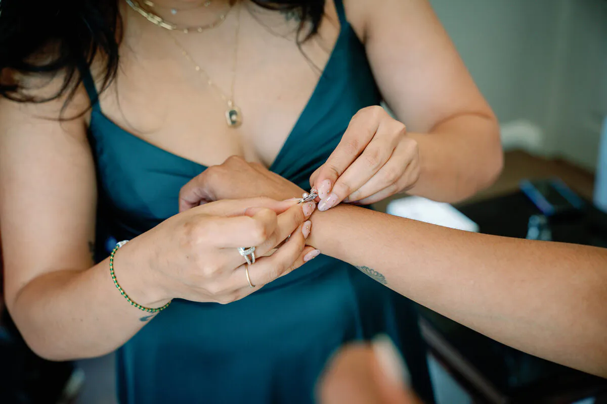 A bridesmaid wearing a dark green silk dress fastens a delicate bracelet onto another person's wrist during wedding preparations.