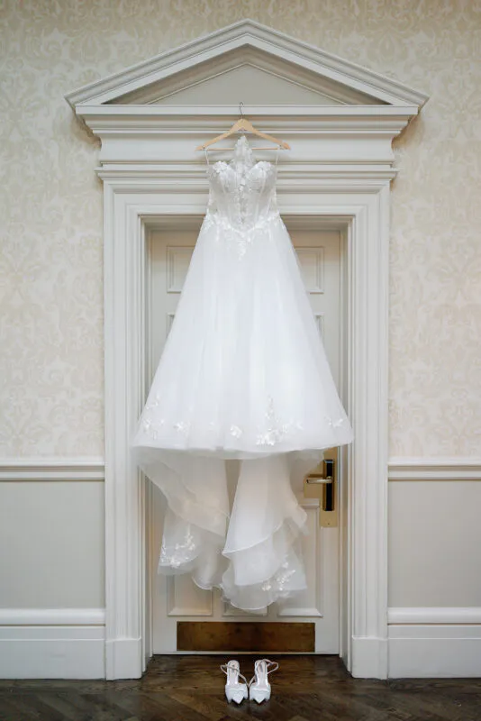 An elegant strapless wedding dress with floral applique hangs from a wooden hanger against a white doorway, with bridal heels placed neatly on the wooden floor below.