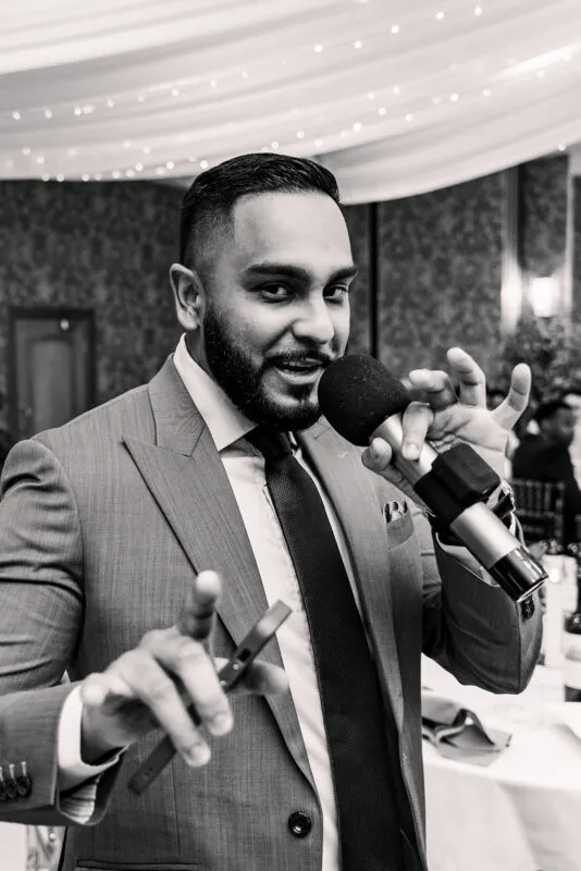 A black and white candid photograph of a groom laughing and gesturing while speaking into a microphone during wedding reception speeches.