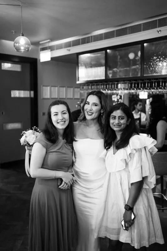 Three guests smile together inside a venue during a wedding celebration, captured in a monochrome documentary style.