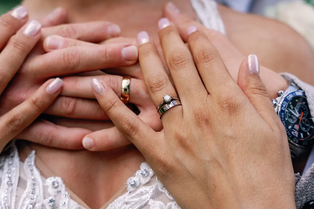 A close-up shot captures the newlyweds embracing, showcasing their stacked wedding rings, engagement diamond, and manicured hands against an intricate lace dress.