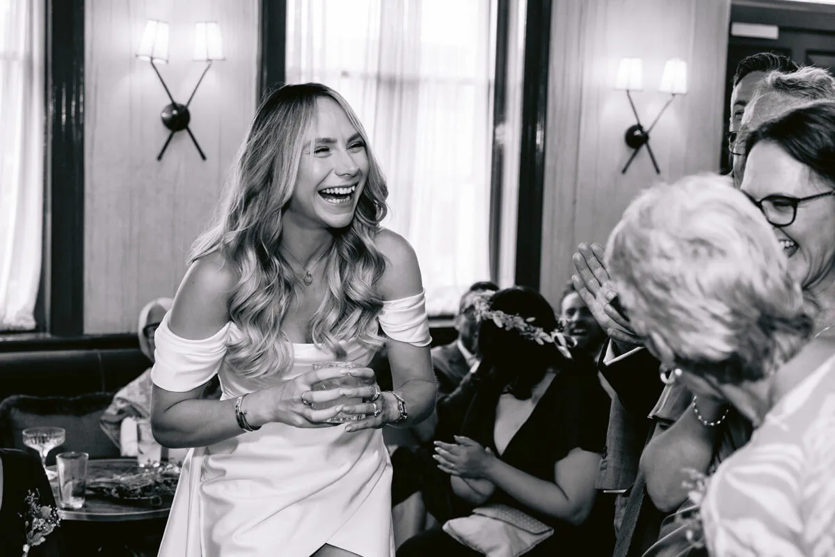 A joyful bride in an off-the-shoulder satin gown laughs heartily while holding a glass at her wedding reception.