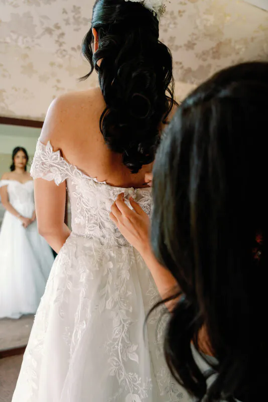 A bridesmaid fastens the back of an off-the-shoulder lace wedding dress as the bride looks into a mirror during bridal preparations.