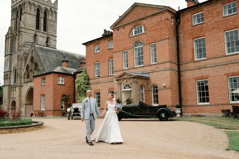 A newlywed couple walk hand in hand across a gravel driveway in front of a grand red brick country house and historic stone church.