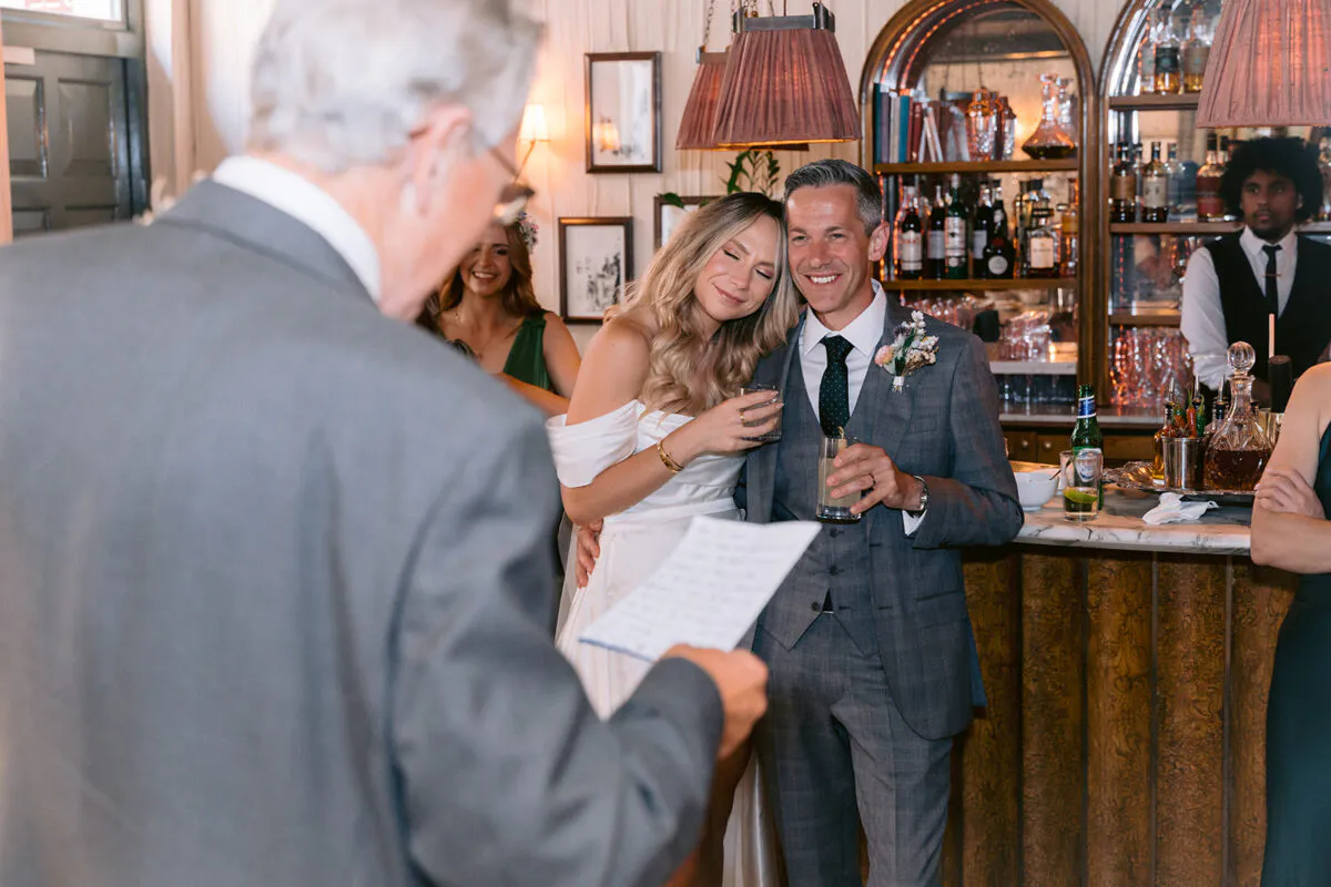 The bride and groom smile as they lean against a pub bar while a guest reads a speech in the foreground.