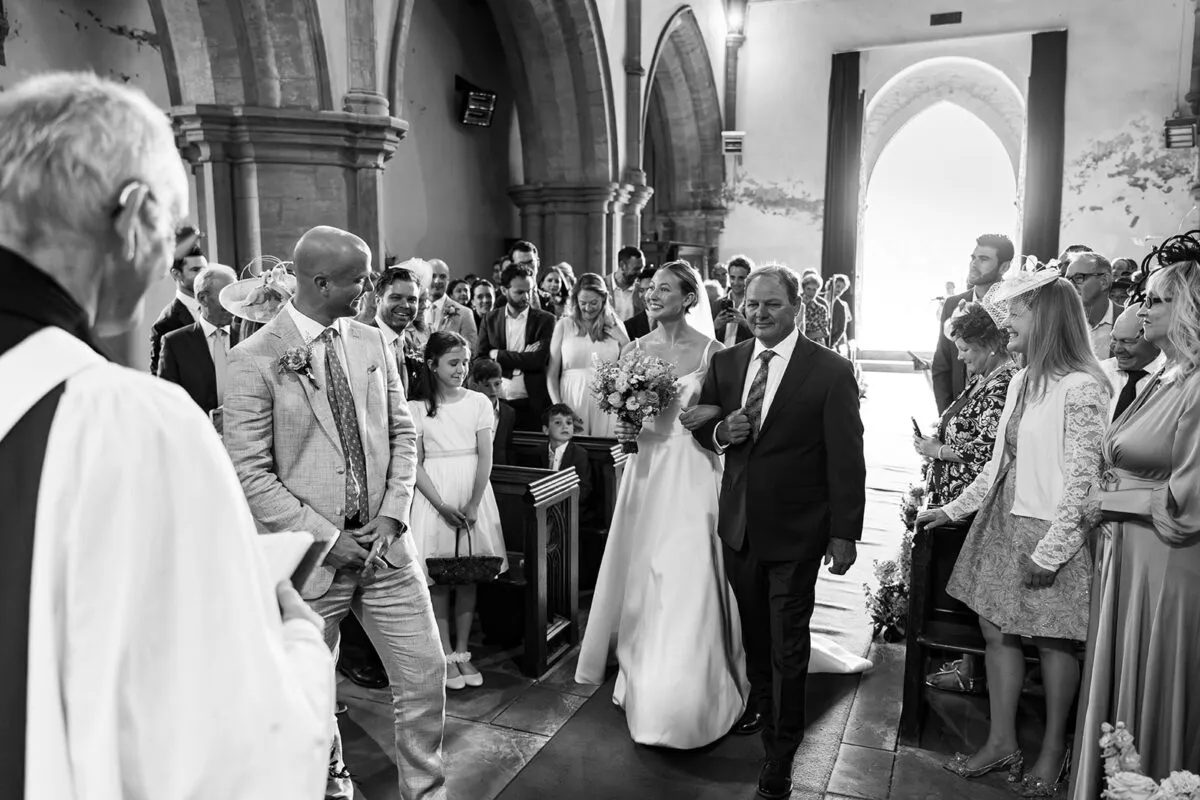 The bride walks down the aisle accompanied by her father inside a historic stone church, smiling towards the groom who waits at the altar.