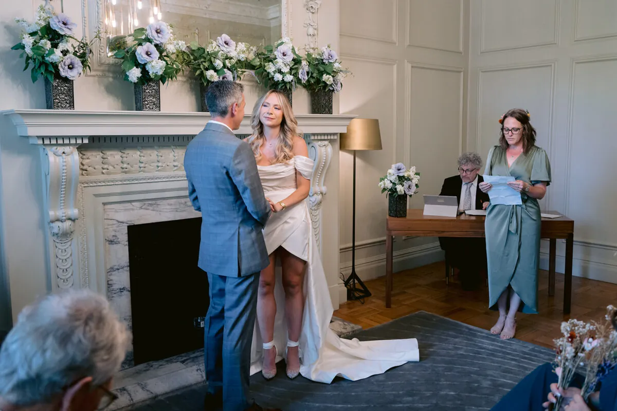 A bride in a modern satin high-low gown and groom in a grey suit stand together during their civil ceremony in a wood-panelled room with a decorated fireplace.