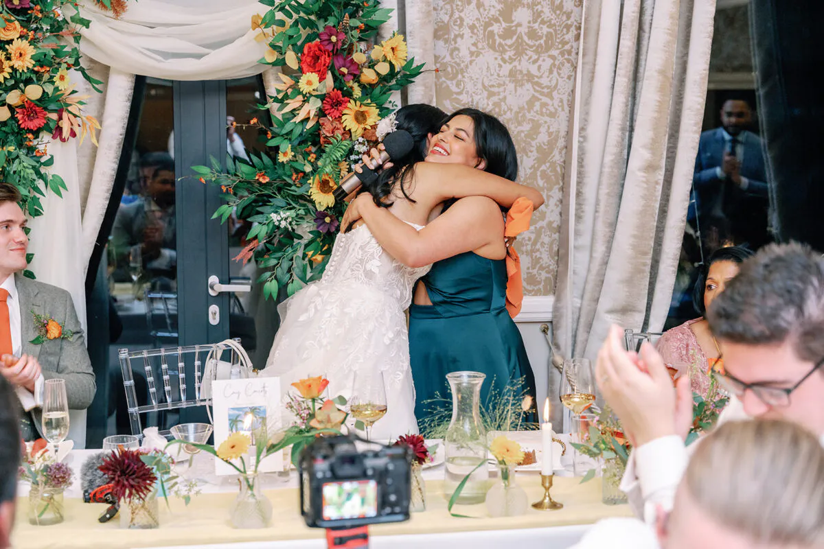 A bride in a lace wedding dress shares an emotional hug with a bridesmaid during the wedding speeches at a beautifully decorated reception table.