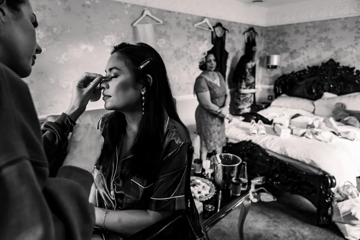 A bride having her makeup applied by a professional while wearing silk pyjamas in a hotel room with family members visible in the background.