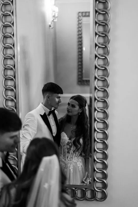A bride and groom share a tender glance reflected in a large, ornately framed mirror.