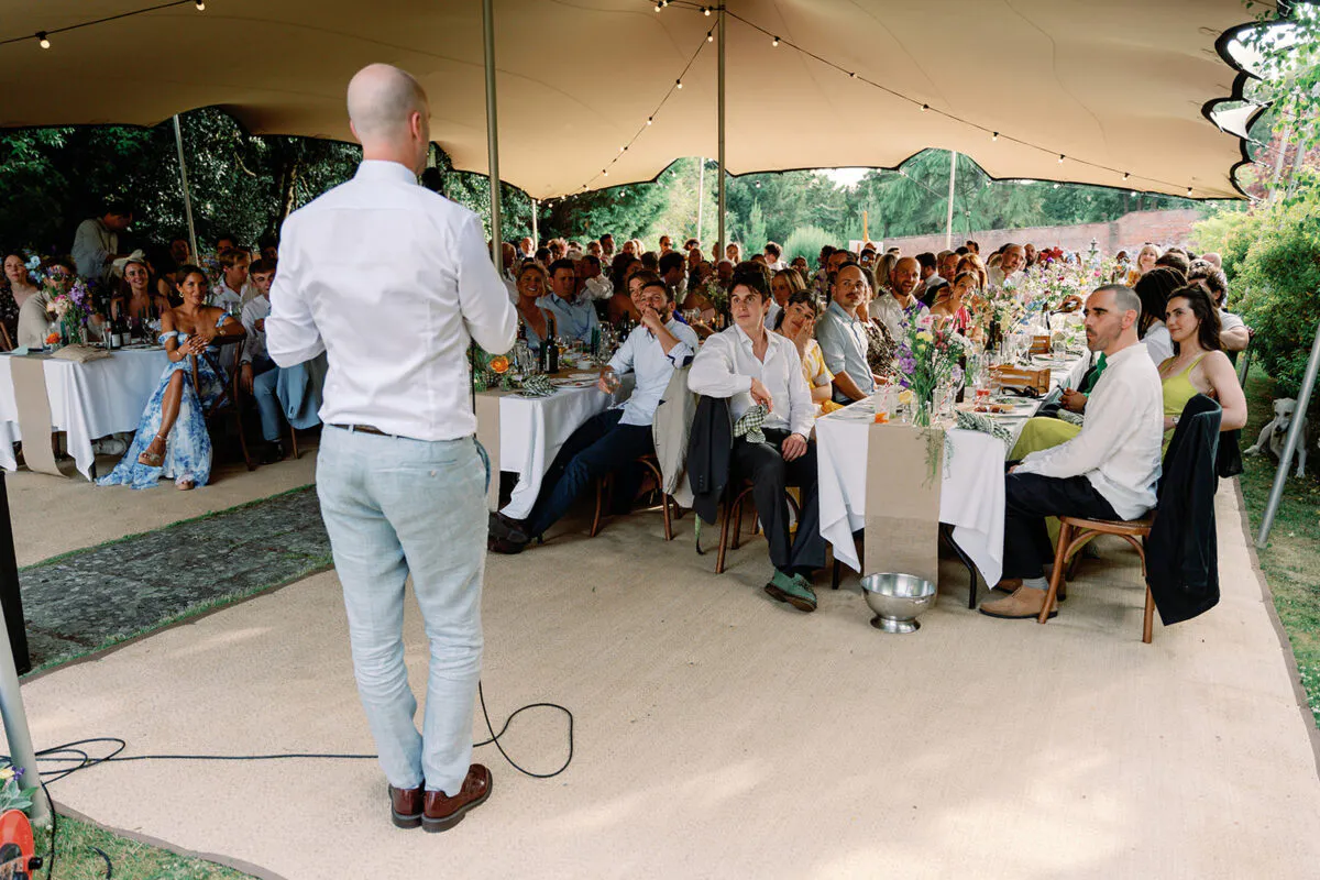 A groom stands with a microphone, delivering a speech to seated guests under a large stretch tent at an outdoor wedding reception.