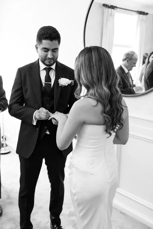 A groom in a dark three-piece suit carefully places a ring onto the finger of his bride, who wears a strapless white satin gown with her hair styled in soft waves.