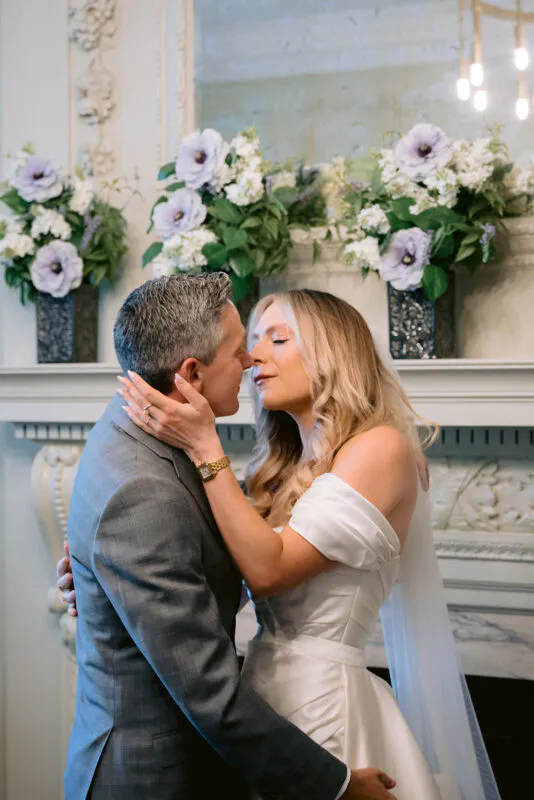 A bride and groom share a tender embrace near a decorated fireplace featuring purple floral arrangements.