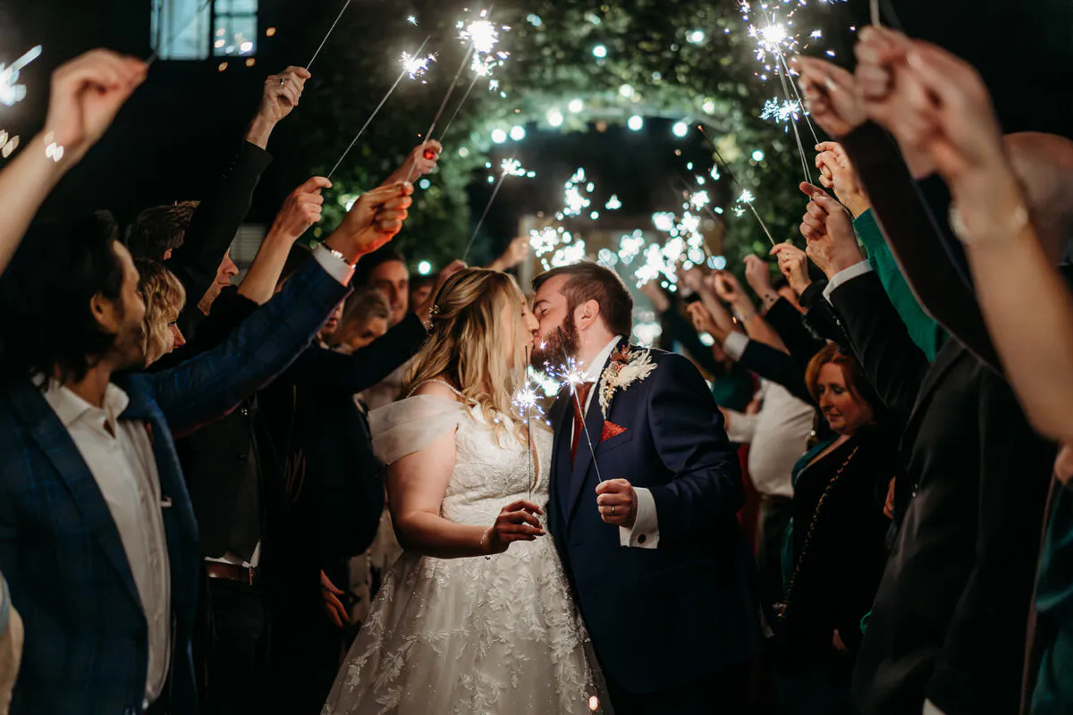A newlywed couple shares a romantic kiss as they walk through a glowing sparkler tunnel held by cheering guests at night.
