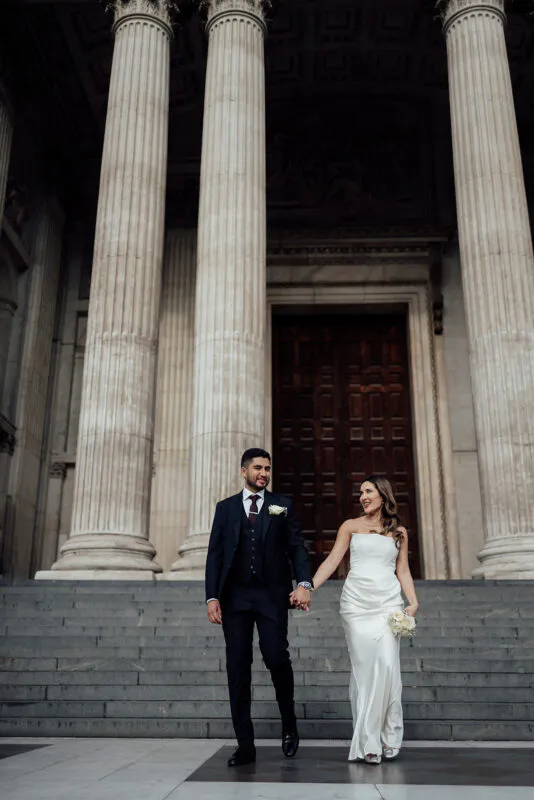A bride and groom holding hands and walking down grand stone steps in front of towering architectural columns at a city wedding.