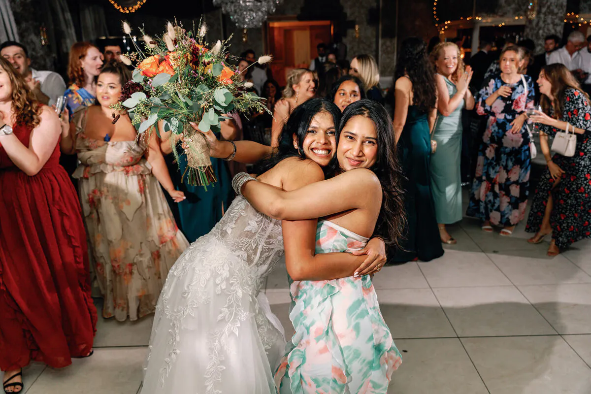 A smiling bride in a lace gown embraces a guest tightly on a lively evening dance floor while holding a large, unstructured bouquet.