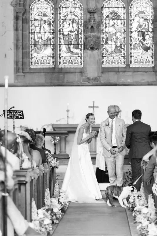 A joyous newlywed couple laughs happily while walking down the church aisle together, accompanied by a small dog on a leash amidst floral arrangements and seated guests.