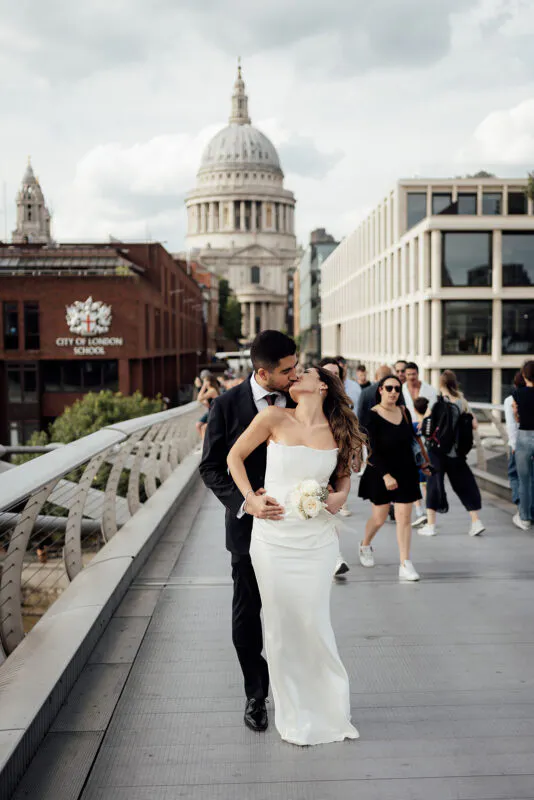 A newlywed couple shares a romantic kiss on a bustling London bridge with St Paul's Cathedral rising in the background.