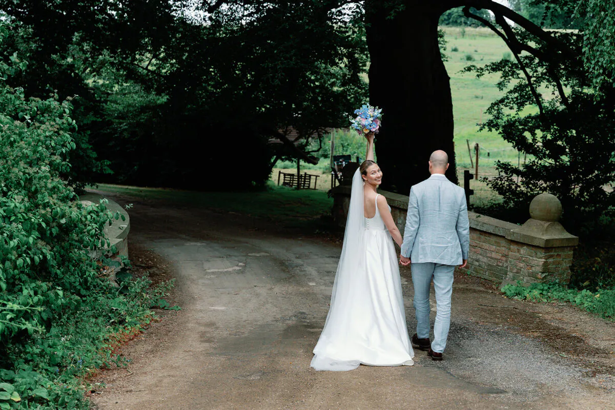 A bride and groom walk hand in hand along a country pathway near a stone bridge, with the bride looking back over her shoulder and holding up a bouquet.
