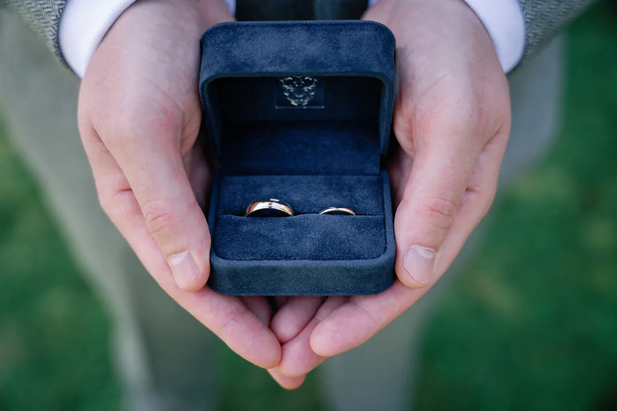 A close-up view of hands gently holding an open blue velvet ring box containing a pair of gleaming wedding bands.