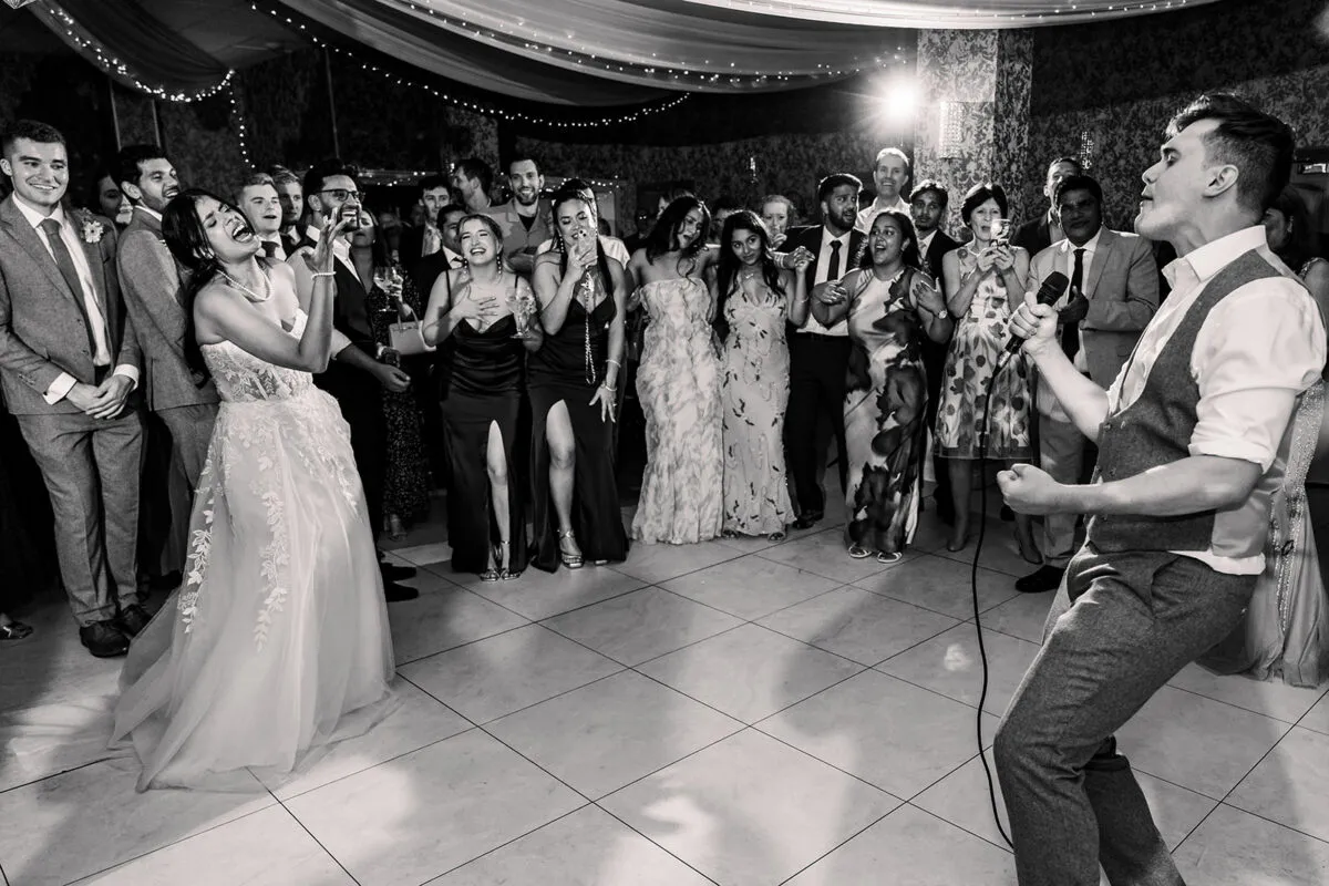 A joyful bride and a singer perform on a packed dance floor while wedding guests cheer and laugh in the background during an evening reception.