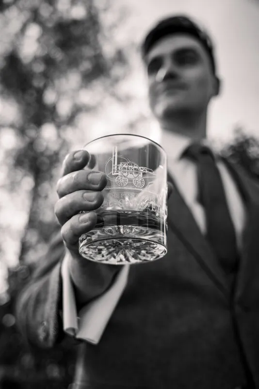 A sharp, low-angle black and white shot of a groom in a suit holding up an engraved whisky tumbler towards the camera.
