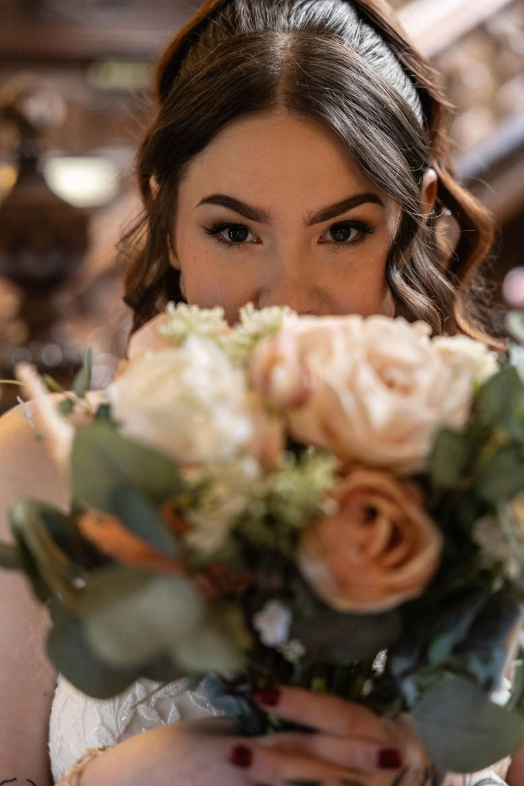 A beautiful bride holds a lush hand-tied rose bouquet in front of her face, peering over the flowers with soft glam makeup and half-up wavy hair.