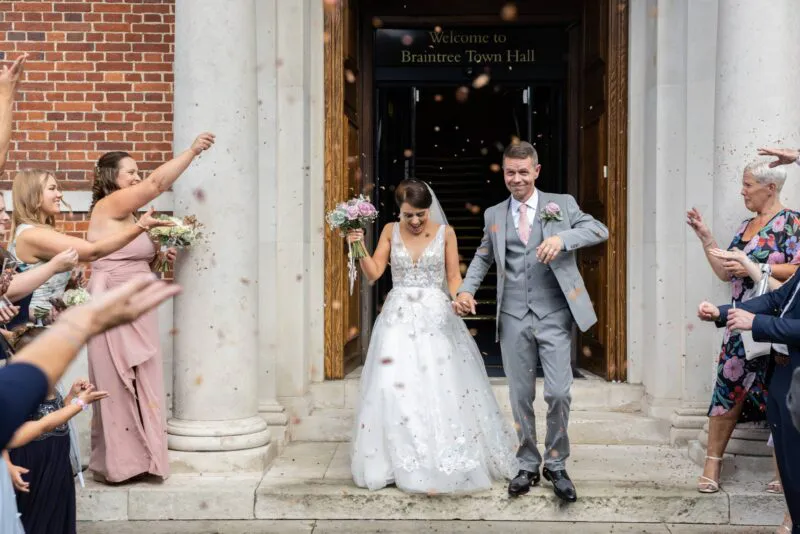 A newlywed couple joyfully exits Braintree Town Hall holding hands while guests shower them with confetti.