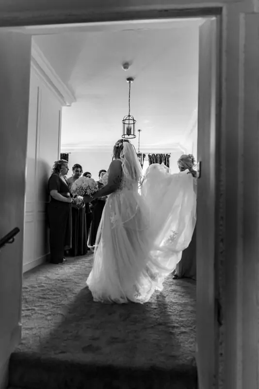 A bride in a lace and tulle wedding gown twirls her dramatic dress and veil in a light-filled room while bridesmaids watch from the side, captured through an open doorway in a striking black and white photograph.