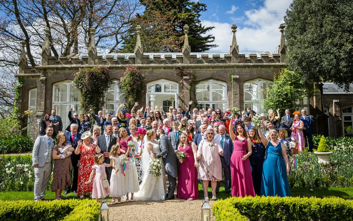 A large group of wedding guests and the bridal party cheer and raise their arms in celebration outside a historic stone orangery on a bright spring day.