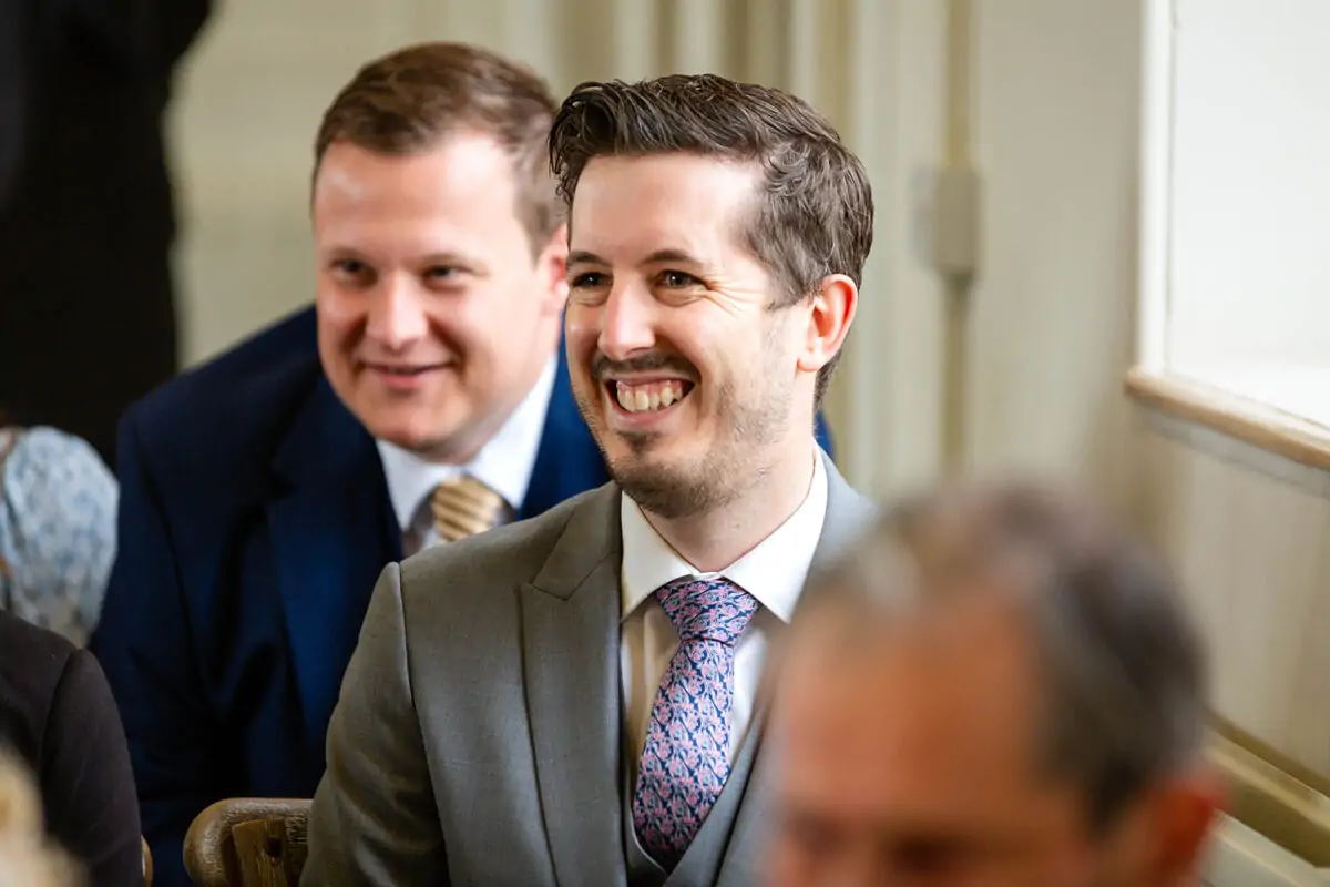 A smiling guest in a grey suit and patterned tie shares a joyful moment during the wedding ceremony.